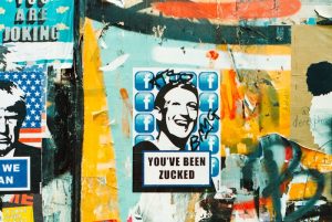 A collage of street art posters on a colourful, weathered wall. The central poster features a stylized black-and-white image of a a smiling Mark Zuckerberg, over a background of Facebook logos, with the text 'YOU'VE BEEN ZUCKED' below. The poster is defaced with graffiti. The surrounding area is filled with layers of torn posters, paint, and scribbles.